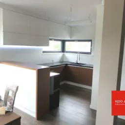 Nowoczesna kuchnia z białymi szafkami górnymi podświetlonymi LED, drewnianymi blatami i brązowymi szafkami dolnymi, widok na okno i zlewozmywak, częściowo widoczny stół z krzesłami w folii...