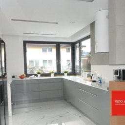 KĘDO Kędzior Dariusz - Nowoczesna, jasna kuchnia z szarymi szafkami, białym blatem i marmurową podłogą, widok na ogród przez duże okno, zlew pod oknem, okrągły biały okap, na blacie owoce i kwiaty w doniczkach, z prawej...