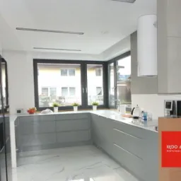 Nowoczesna, jasna kuchnia z szarymi szafkami, białym blatem i marmurową podłogą, widok na ogród przez duże okno, zlew pod oknem, okrągły biały okap, na blacie owoce i kwiaty w doniczkach, z prawej...