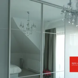 Szafa przesuwna z lustrzanymi drzwiami, odbijająca wnętrze pokoju z żyrandolem kryształowym i łóżkiem, z widocznym logo producenta mebli.