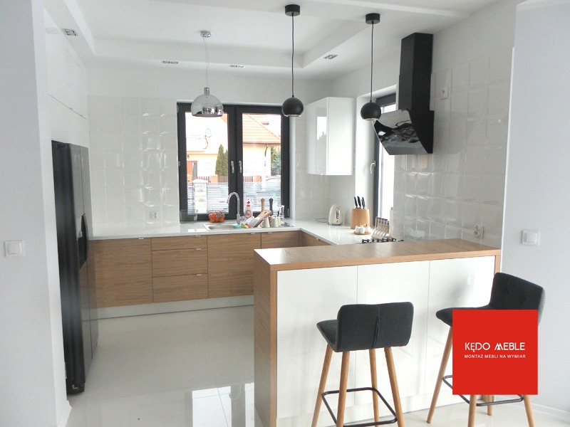 Nowoczesna kuchnia z białymi szafkami wiszącymi, drewnianymi frontami szafek dolnych i wyspą z dwoma hokerami, czarnym okapem i błyszczącymi płytkami na ścianie.