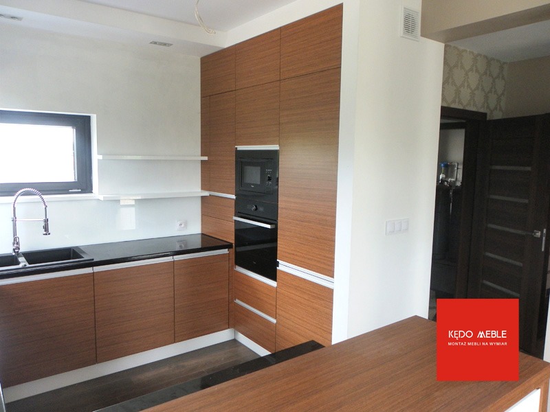 Nowoczesna kuchnia z drewnianymi frontami szafek, czarnym blatem i wbudowanym sprzętem AGD, widoczny fragment wyspy kuchennej i logo firmy meblarskiej w prawym dolnym rogu.
