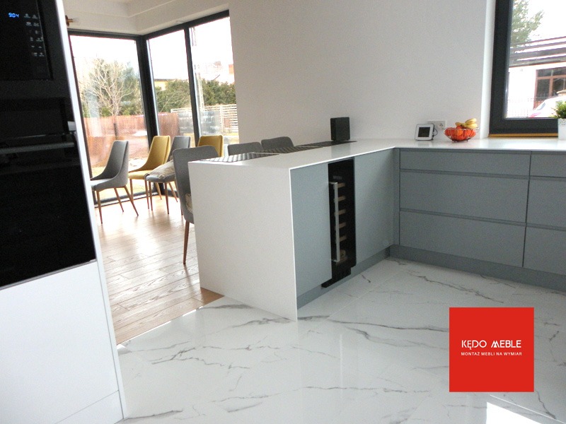 Nowoczesna kuchnia z szarymi szafkami, białą wyspą i podłogą z marmuru, wbudowana chłodziarka na wino, widok na ogród przez duże okna.