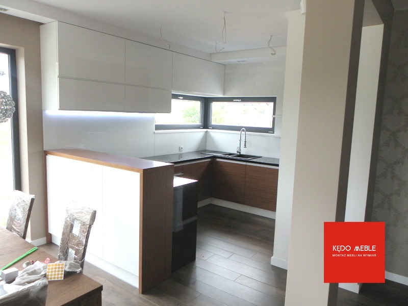 Nowoczesna kuchnia z białymi szafkami górnymi podświetlonymi LED, drewnianymi blatami i brązowymi szafkami dolnymi, widok na okno i zlewozmywak, częściowo widoczny stół z krzesłami w folii...