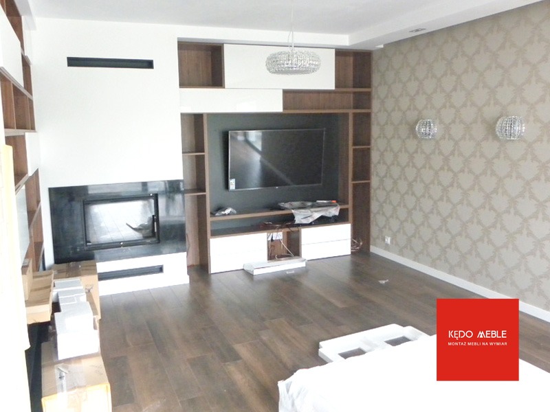 Salon z kominkiem i zabudową meblową w trakcie montażu, widoczne kartony i elementy wykończeniowe, parkiet ułożony w jodełkę, ściana w ozdobną tapetę.