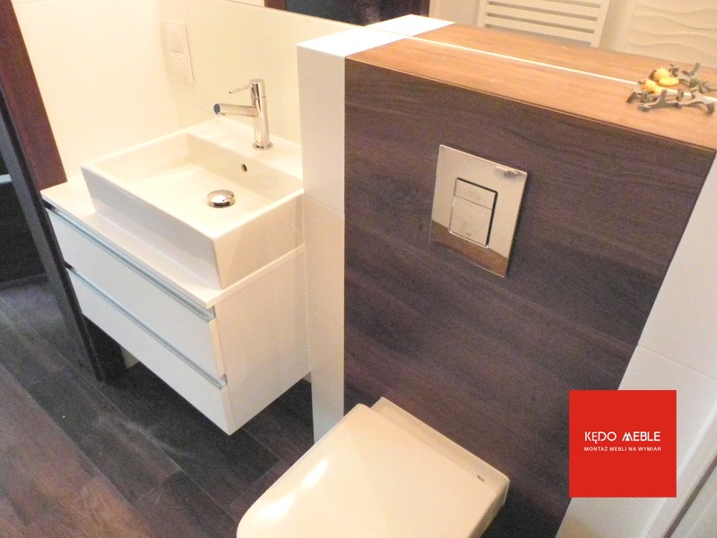 Nowoczesna łazienka z białą umywalką nablatową na szafce z szufladami, chromowaną baterią i wiszącą muszlą klozetową z panelem w kolorze ciemnego drewna.