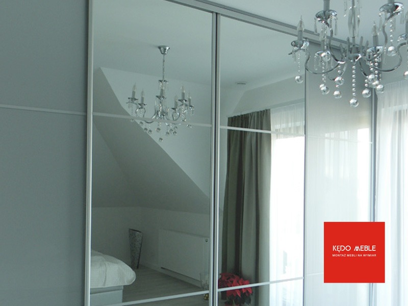 Szafa przesuwna z lustrzanymi drzwiami, odbijająca wnętrze pokoju z żyrandolem kryształowym i łóżkiem, z widocznym logo producenta mebli.