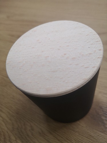 Czarne, matowe pudełko z jasną, drewnianą pokrywką, ustawione ukośnie na drewnianej powierzchni.