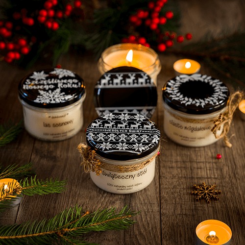 Cztery słoiczki z naturalnymi świecami sojowymi o zapachach lasu sosnowego, ciasta migdałowego i świątecznym motywem, ozdobione sznurkiem jutowym, na drewnianym stole z gałązkami świerku...