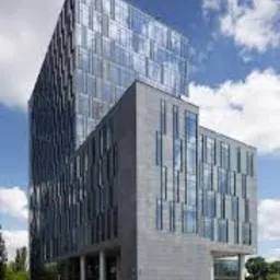 Nowoczesny biurowiec z szklaną fasadą i elementami z szarego kamienia, widoczny z dołu pod błękitnym niebem z chmurami.