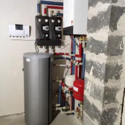 Nowoczesna kotłownia z białym piecem gazowym, szarym zasobnikiem ciepłej wody i czerwoną przeponą, widoczne rury w izolacji niebieskiej i czerwonej, oraz czarne rozdzielacze, na ścianie biały...