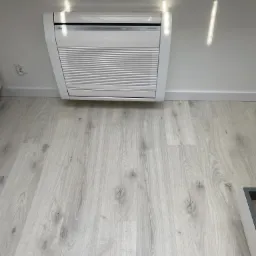 Biała jednostka klimatyzacyjna Fujitsu zamontowana na ścianie nad jasną, drewnianą podłogą, z widocznym gniazdkiem elektrycznym i refleksami światła.