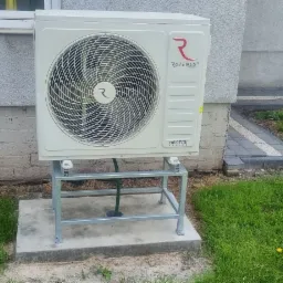 Zewnętrzna jednostka pompy ciepła Rotenso zamontowana na metalowym stelażu przy ścianie budynku z widocznym fragmentem elewacji i trawnikiem.