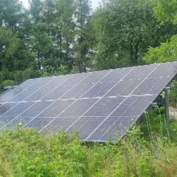 Instalacja paneli słonecznych na metalowej konstrukcji, otoczona bujną zielenią, z widocznymi drzewami w tle i elementami starej zabudowy.