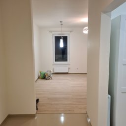 Patryk Kalinowski - Puste, jasne wnętrze po remoncie z drewnianą podłogą, widok z korytarza. Dwa worki stoją pod oknem. Minimalistyczne lampy sufitowe.