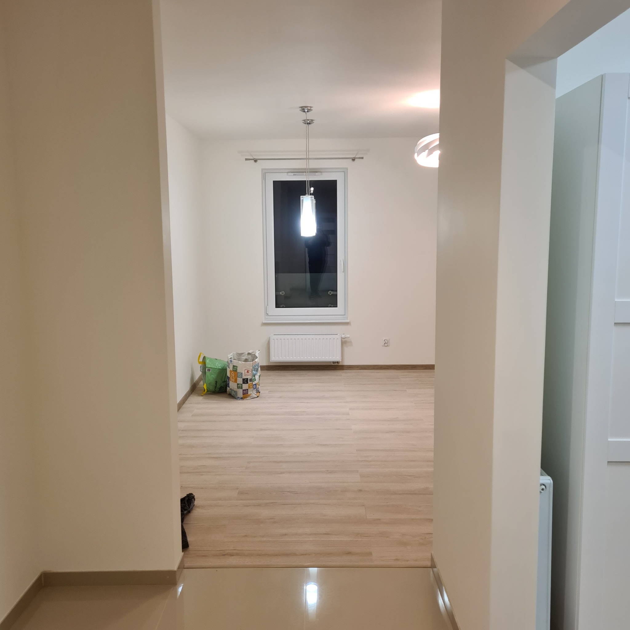 Puste, jasne wnętrze po remoncie z drewnianą podłogą, widok z korytarza. Dwa worki stoją pod oknem. Minimalistyczne lampy sufitowe.