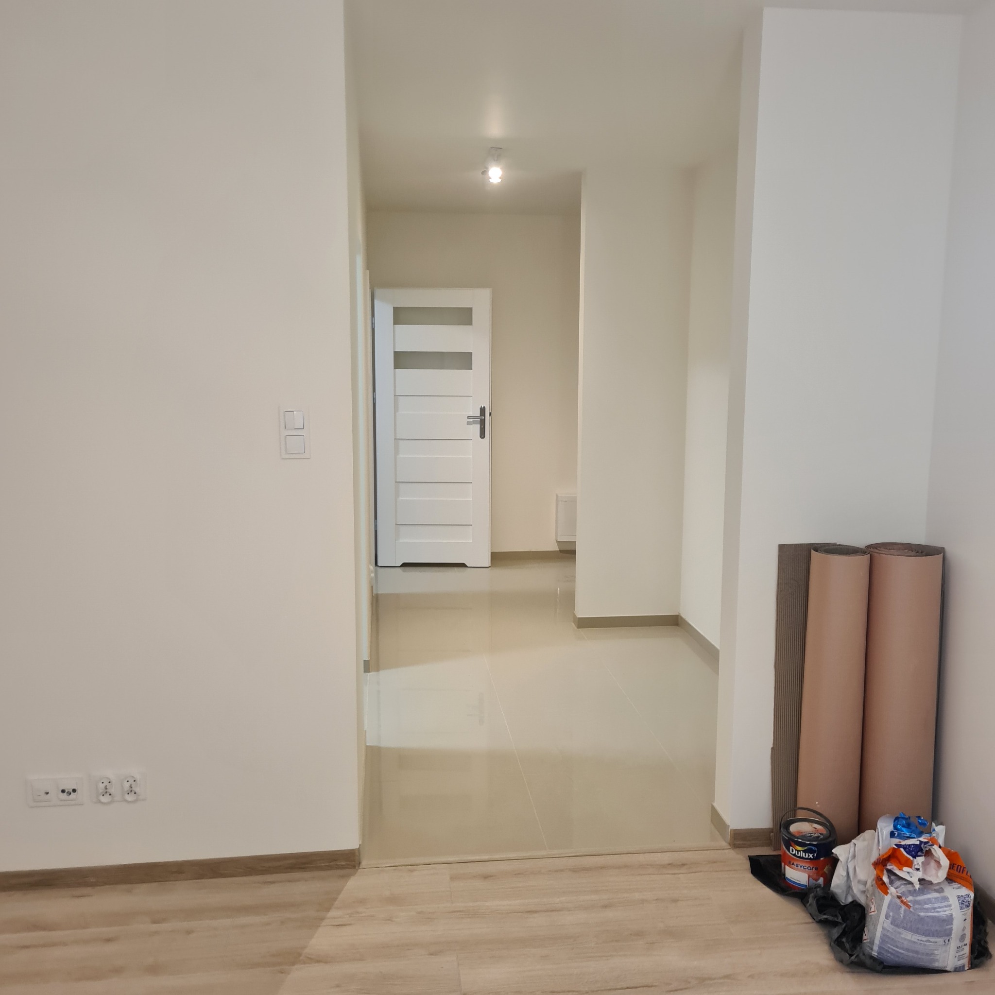 Jasne, odświeżone wnętrze mieszkania po remoncie. Widoczne gładkie, białe ściany, nowe gniazdka elektryczne, drzwi z przeszkleniami i materiały malarskie w rogu.