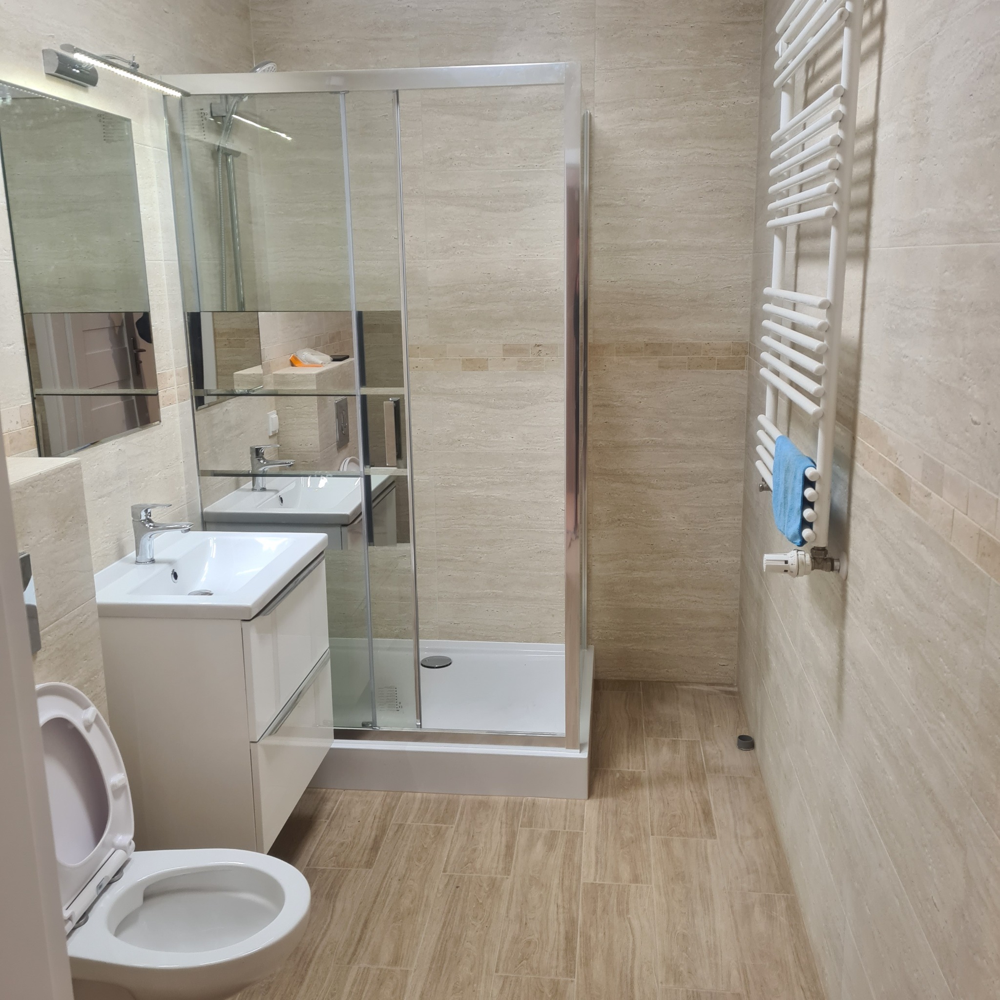 Jasna łazienka z kabiną prysznicową, umywalką z szafką i grzejnikiem drabinkowym z ręcznikiem. Beżowe płytki imitujące trawertyn na ścianach i podłodze.