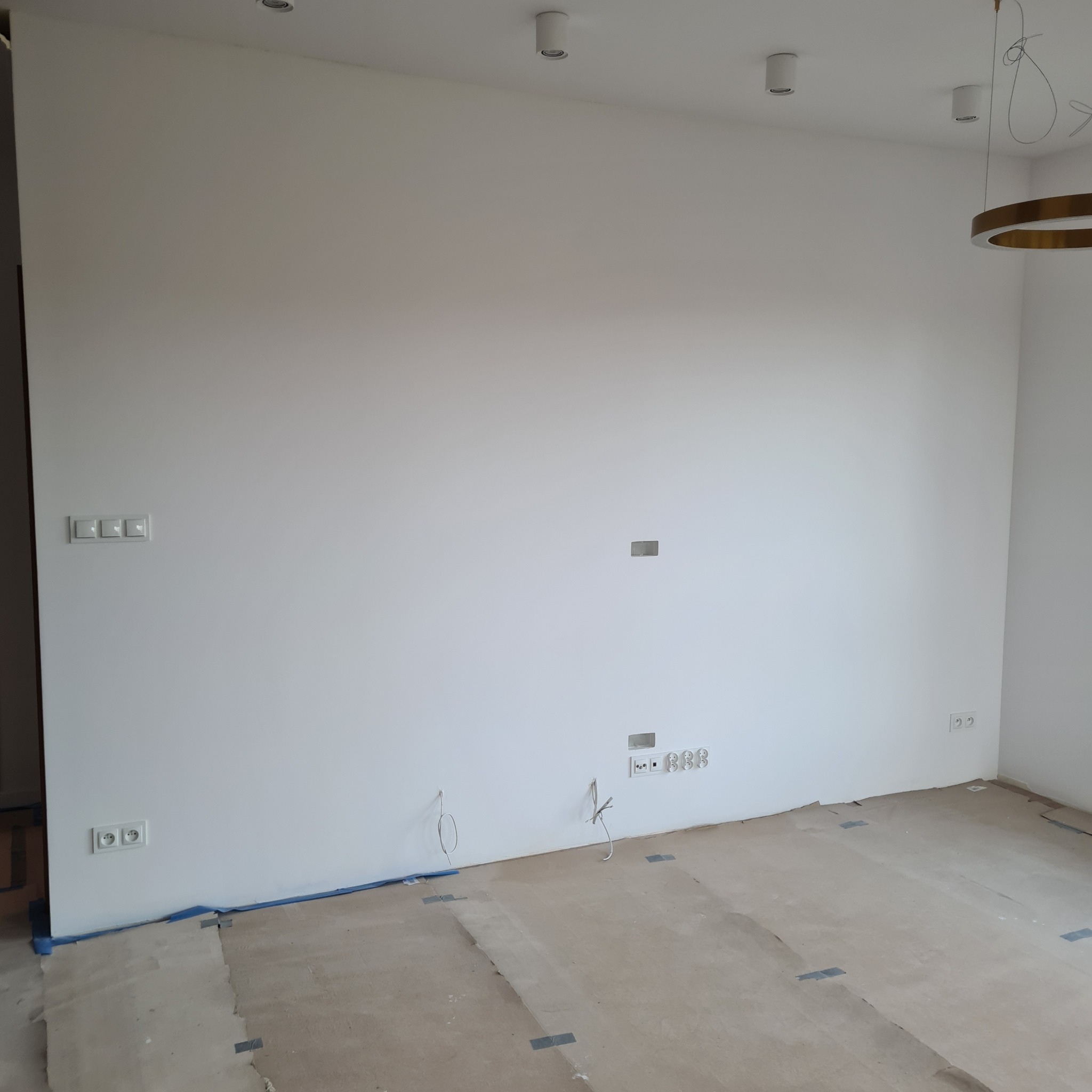 Biała ściana po szpachlowaniu z wystającymi przewodami elektrycznymi i zabezpieczoną podłogą. Widoczne gniazdka i włączniki. Przygotowanie do wykończenia wnętrz.
