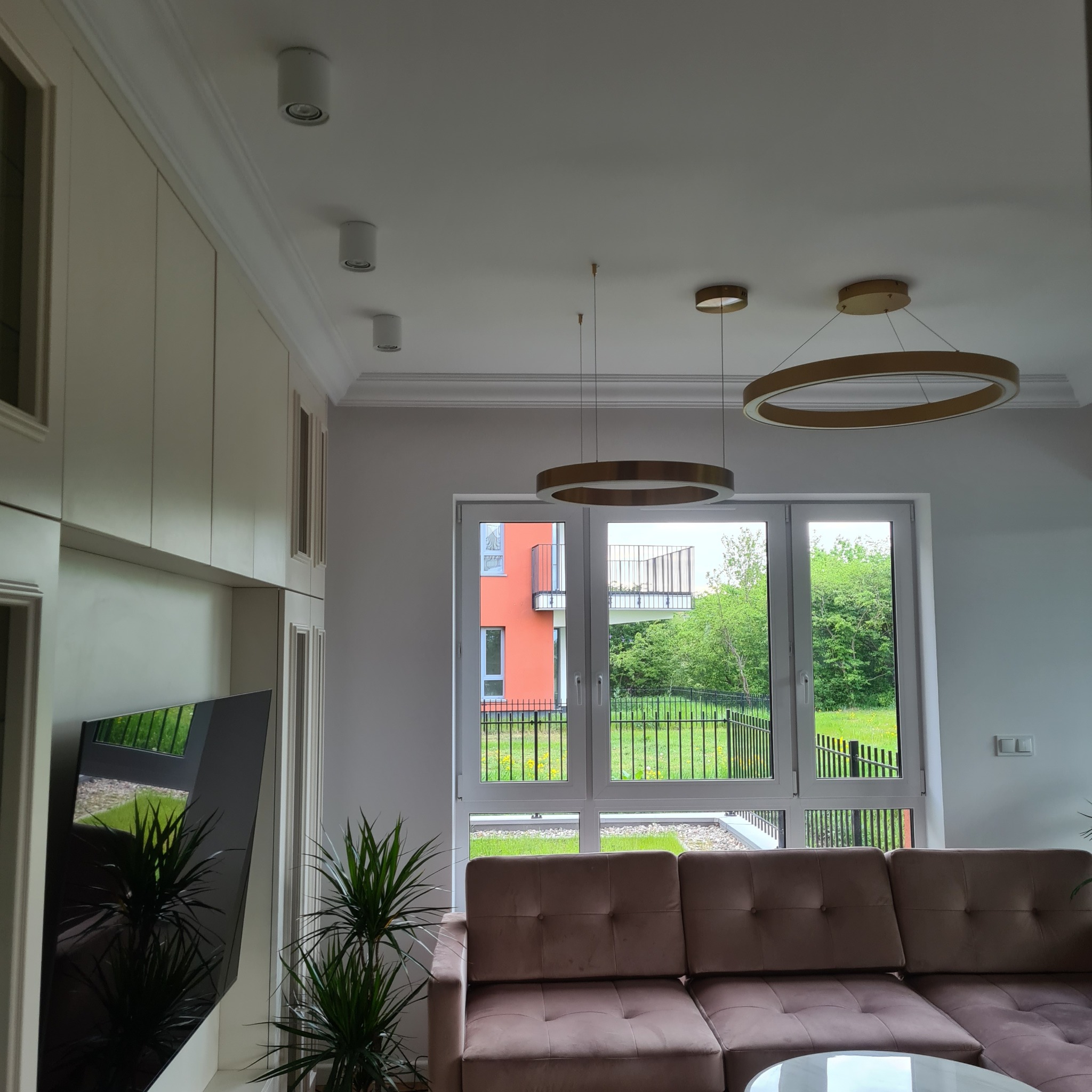 Nowoczesny salon z beżową sofą, okrągłymi lampami i widokiem na zieleń za oknem. Minimalistyczny design, jasne kolory i rośliny doniczkowe tworzą przytulną atmosferę.