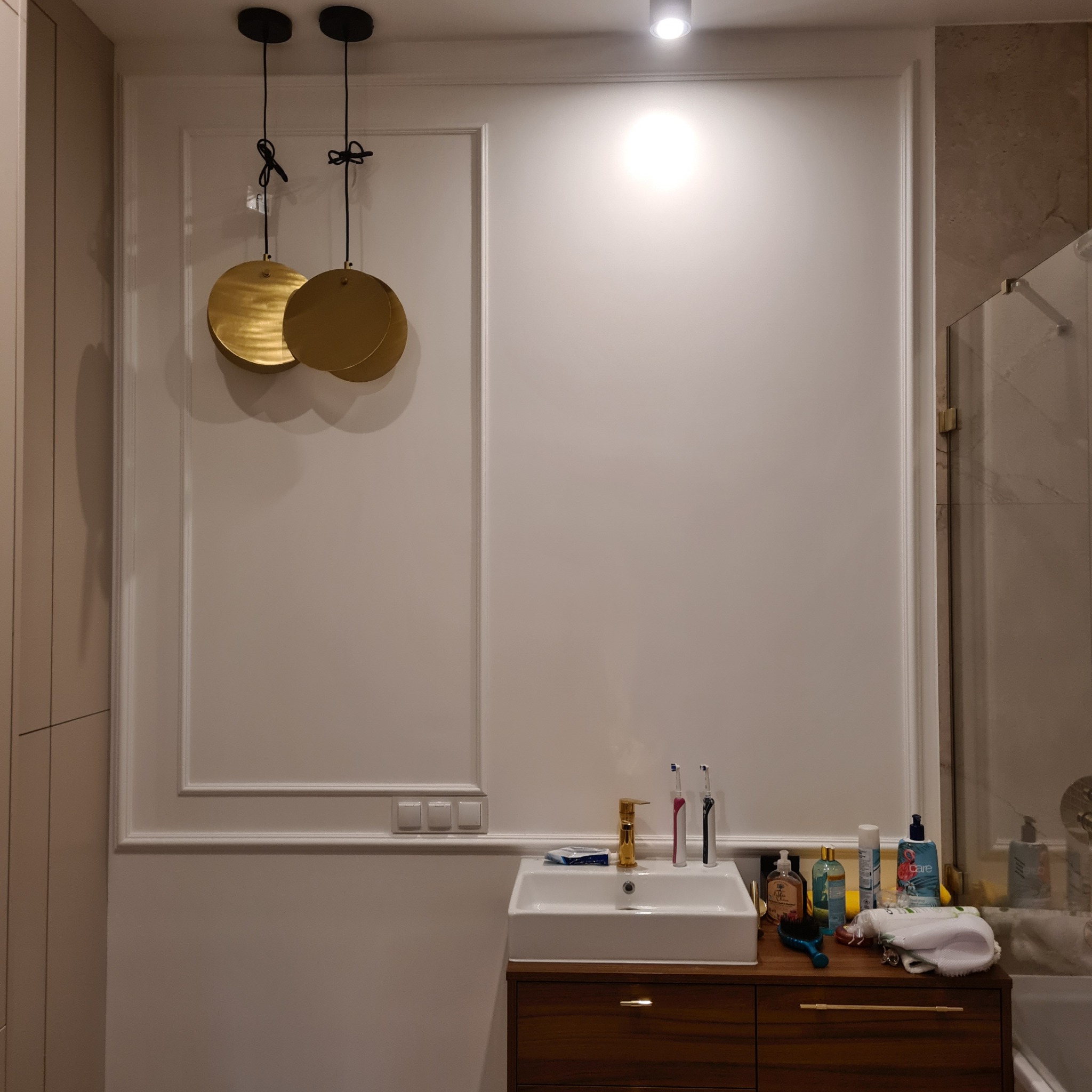Wykończona łazienka z białą umywalką na drewnianej szafce, złotą armaturą i okrągłymi, złotymi lampami wiszącymi. Ściany zdobią białe listwy dekoracyjne.