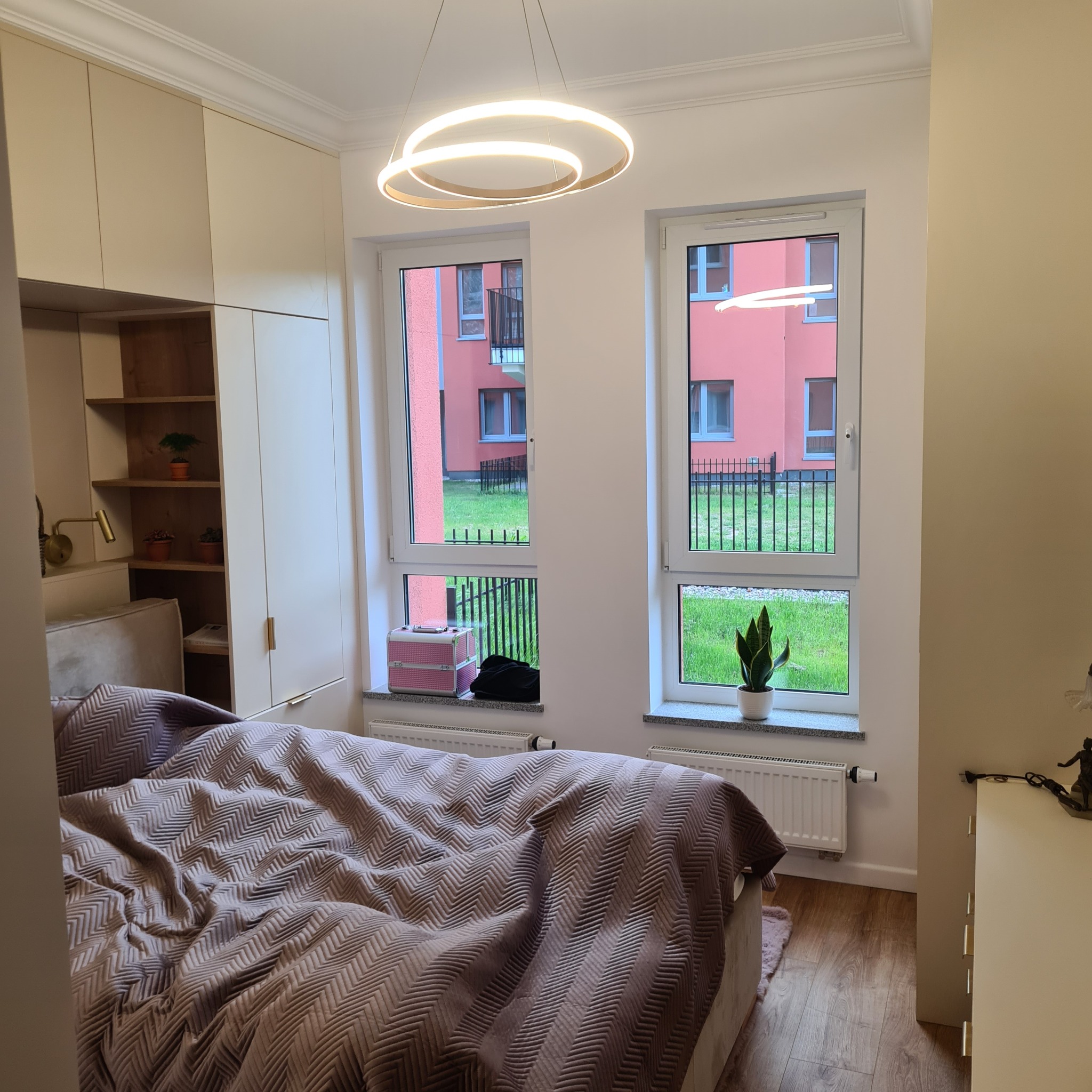 Jasna sypialnia z łóżkiem w zygzaki, okrągłą lampą i widokiem na zieleń. Na parapecie różowa walizka i roślina w doniczce. Nowoczesne wykończenie wnętrza.