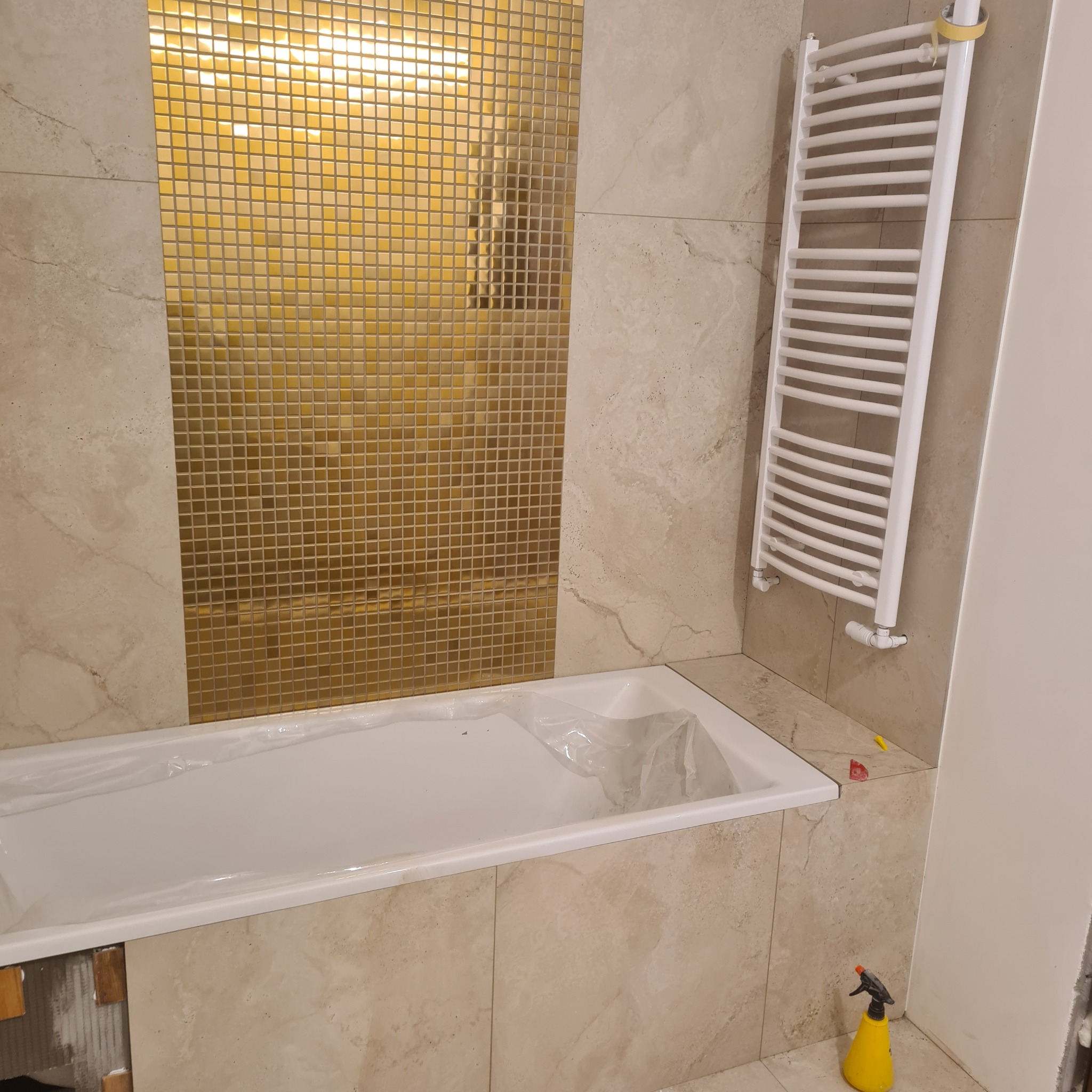 Wykończona łazienka z wanną zabezpieczoną folią, grzejnikiem drabinkowym i złotą mozaiką na ścianie. Widoczny spryskiwacz i elementy montażowe płytek.