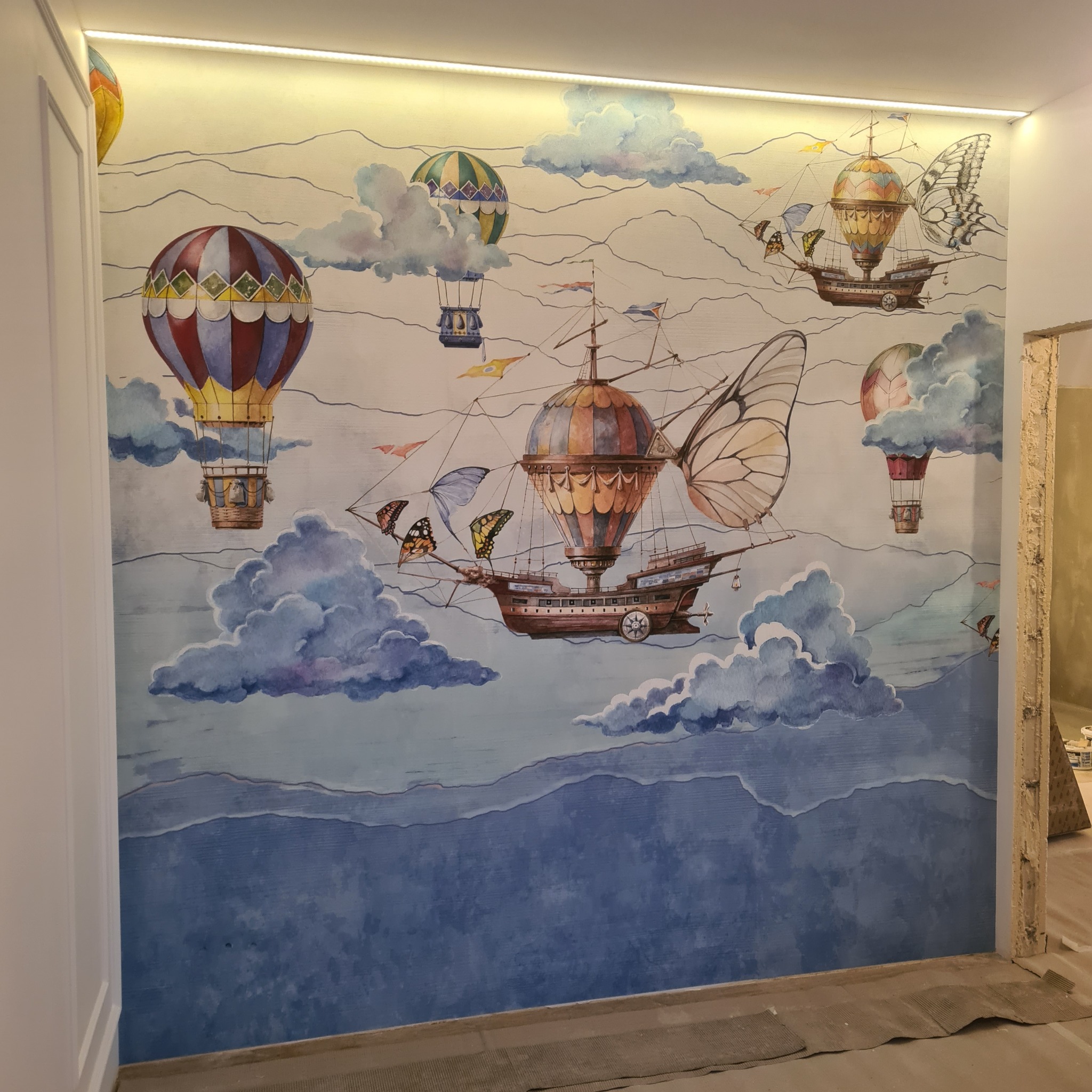 Ściana z ręcznie malowanym, fantazyjnym motywem: balony, statki powietrzne z motylimi skrzydłami, chmury i góry w odcieniach błękitu. Oświetlenie LED u góry.