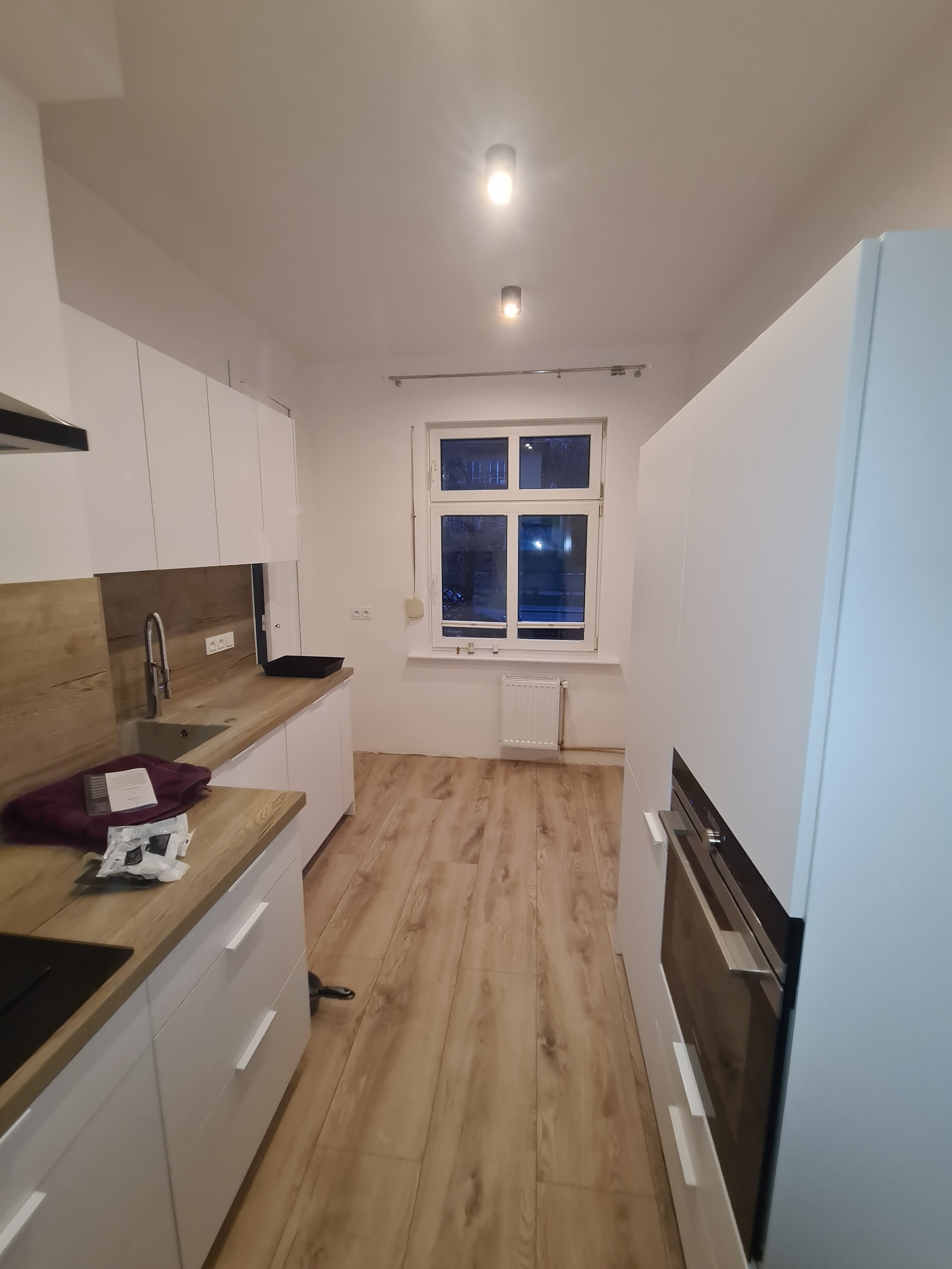 Nowoczesna kuchnia z białymi szafkami i drewnianym blatem, widok na okno. Podłoga wyłożona panelami imitującymi drewno. Oświetlenie sufitowe.