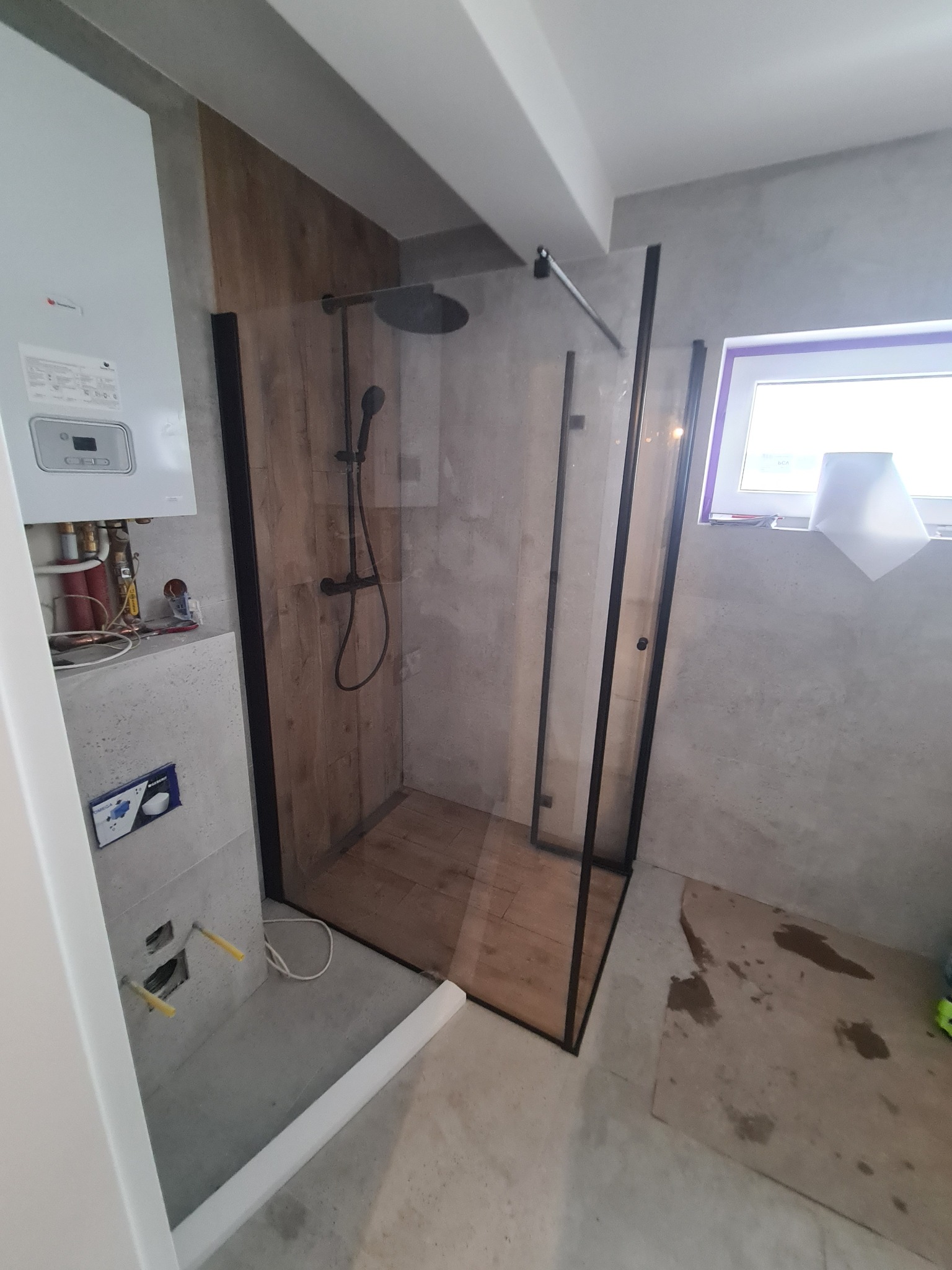 Nowoczesna kabina prysznicowa z czarnymi ramami i drewnianym wykończeniem wewnątrz, zamontowana w łazience z szarą ścianą i oknem. Widoczny bojler i instalacja.