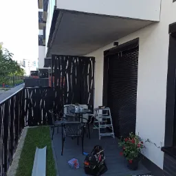 Zadaszony balkon w nowoczesnym budownictwie z czarnymi roletami zewnętrznymi, ażurową balustradą i zestawem mebli ogrodowych, na podłodze rozłożone narzędzia i drabina.