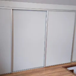 Czterodrzwiowa szafa wnękowa z przesuwanymi, jasnoszarymi drzwiami, wkomponowana w przestrzeń pod schodami, z widocznym fragmentem drewnianej podłogi i futryny drzwi.