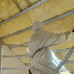 Pracownik w kombinezonie ochronnym na rusztowaniu montuje izolację z wełny mineralnej na poddaszu, widoczne drewniane belki stropowe i metalowe profile.