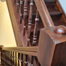 Ciemne, drewniane schody z toczonymi balustradami i poręczą, widoczne z dołu, z wyraźnym usłojeniem drewna i detalami wykończenia.