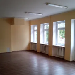 Pusta sala z trzema białymi oknami, brązową podłogą imitującą drewno i żółtymi ścianami, oświetlona lampami sufitowymi.