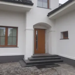 Biała elewacja domu z drewnianą stolarką okienną i wejściową, schody z szarego kamienia, kostka brukowa na podjeździe.