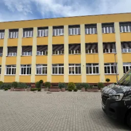 Żółty budynek z białymi oknami i brązowymi roletami zewnętrznymi, widoczny z częściowo zasłaniającego go pojazdu firmy z logo i danymi kontaktowymi.