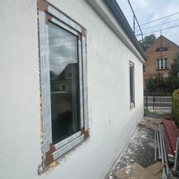 Lubuskie Centrum Okien - Montaż okna PCV marki Salamander w elewacji budynku, widoczna pianka montażowa i folia ochronna na ramie okiennej, w tle dom z cegły i zieleń.