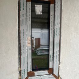 Lubuskie Centrum Okien - Montaż okna z brązową ramą, zabezpieczonego folią ochronną z logo Salamander, w surowej ścianie z widocznymi resztkami pianki montażowej.