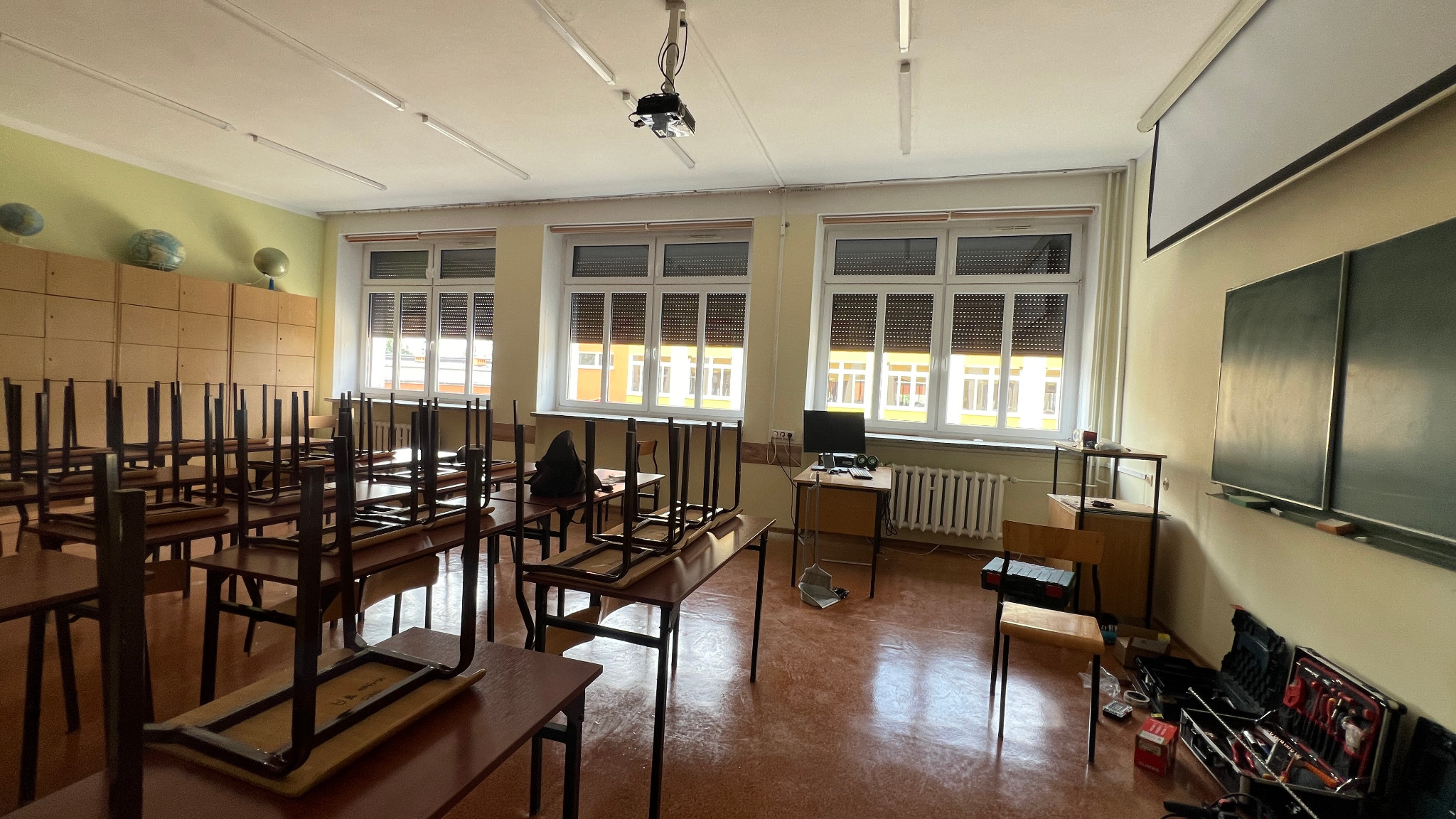 Sala lekcyjna z podniesionymi krzesłami na biurkach, rolety zewnętrzne częściowo zasłonięte w oknach, skrzynka z narzędziami leżąca na podłodze, sugerująca remont lub naprawy.