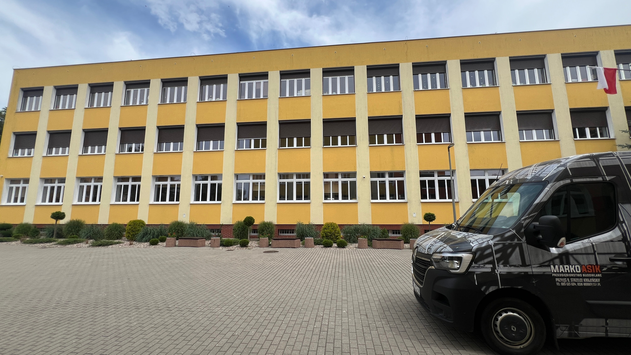 Żółty budynek z białymi oknami i brązowymi roletami zewnętrznymi, widoczny z częściowo zasłaniającego go pojazdu firmy z logo i danymi kontaktowymi.