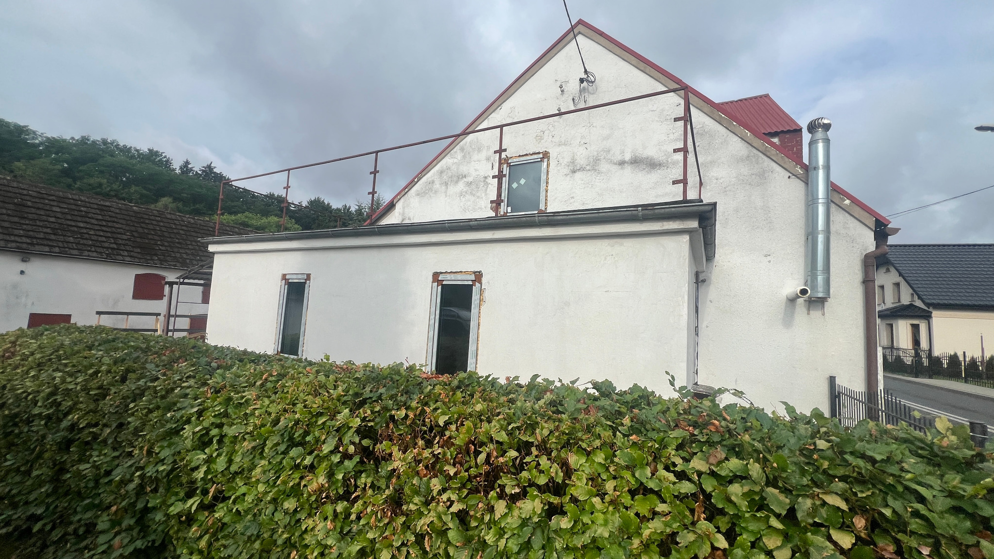 Biały budynek z odkrytymi ramami okiennymi i stalową rurą kominową, widoczny zza wysokiego żywopłotu.