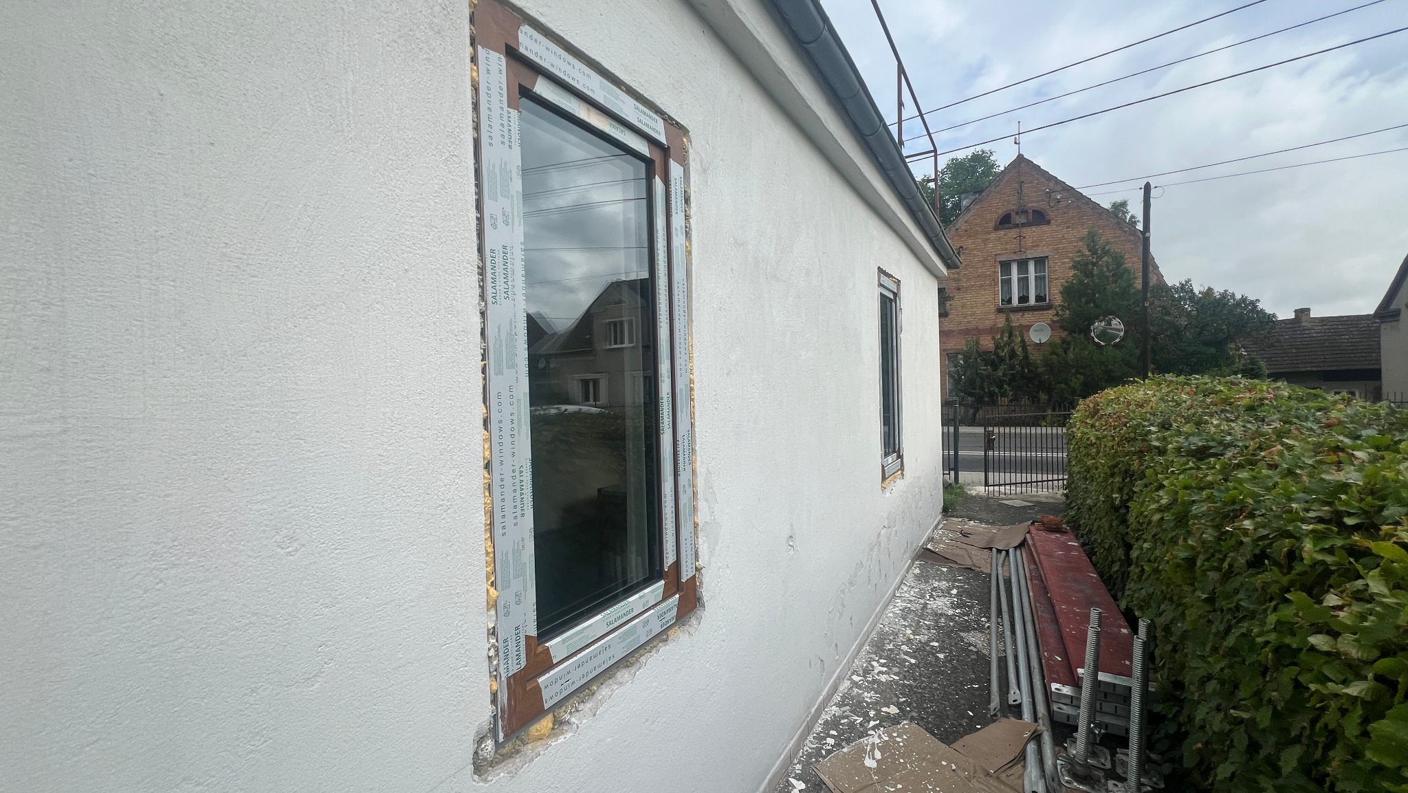 Montaż okna PCV marki Salamander w elewacji budynku, widoczna pianka montażowa i folia ochronna na ramie okiennej, w tle dom z cegły i zieleń.