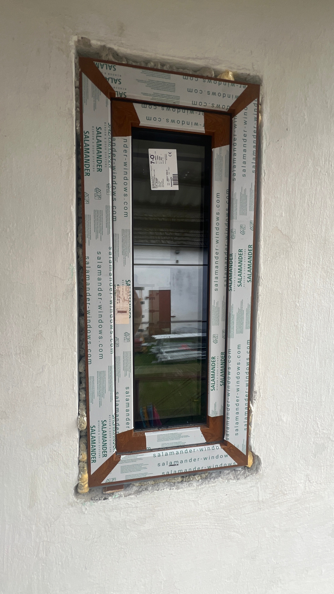 Montaż okna z brązową ramą, zabezpieczonego folią ochronną z logo Salamander, w surowej ścianie z widocznymi resztkami pianki montażowej.