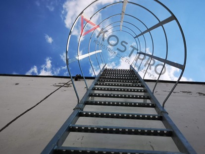 Zewnętrzna drabina ewakuacyjna z metalu, z koszem ochronnym, przymocowana do ściany budynku, widok z dołu na błękitne niebo. Na drabine nałożone jest logo firmy.