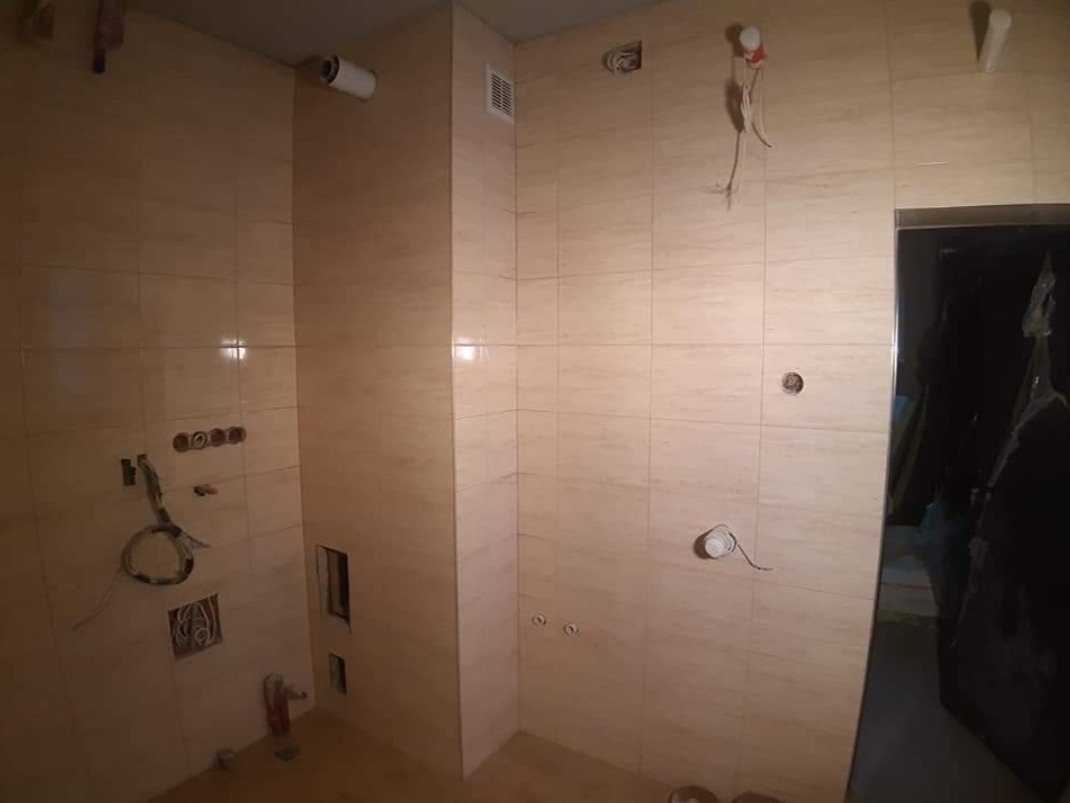 Wnętrze łazienki w trakcie wykańczania, beżowe płytki na ścianach, widoczne przewody elektryczne i instalacje sanitarne przed montażem armatury, otwór wentylacyjny w górnej części ściany.