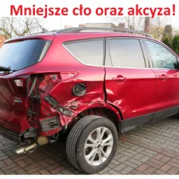 Czerwony SUV z poważnymi uszkodzeniami tylnej części nadwozia, widoczne zgniecenia blachy i odsłonięte elementy konstrukcyjne, na tle brukowanej drogi i zabudowań.