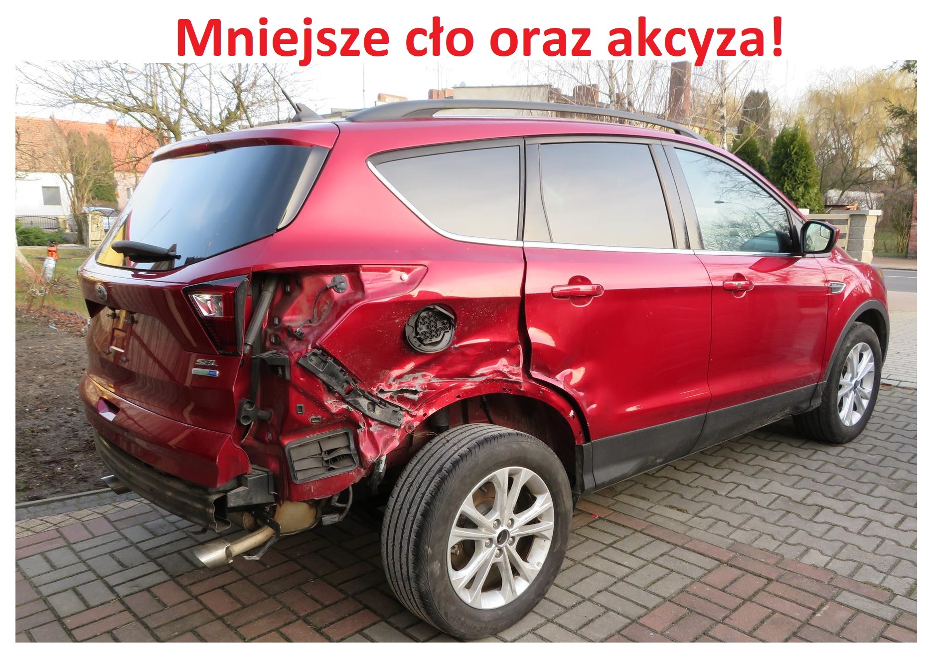 Czerwony SUV z poważnymi uszkodzeniami tylnej części nadwozia, widoczne zgniecenia blachy i odsłonięte elementy konstrukcyjne, na tle brukowanej drogi i zabudowań.