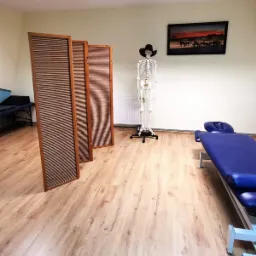 Gabinet z niebieskim łóżkiem do terapii manualnej, parawanem, modelem szkieletu w kapeluszu i aparaturą do fizykoterapii.