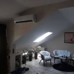 Wnętrze poddasza z zamontowaną jednostką klimatyzacji ściennej, widoczne trzy wiklinowe fotele z poduszkami, stolik kawowy i rośliny doniczkowe.